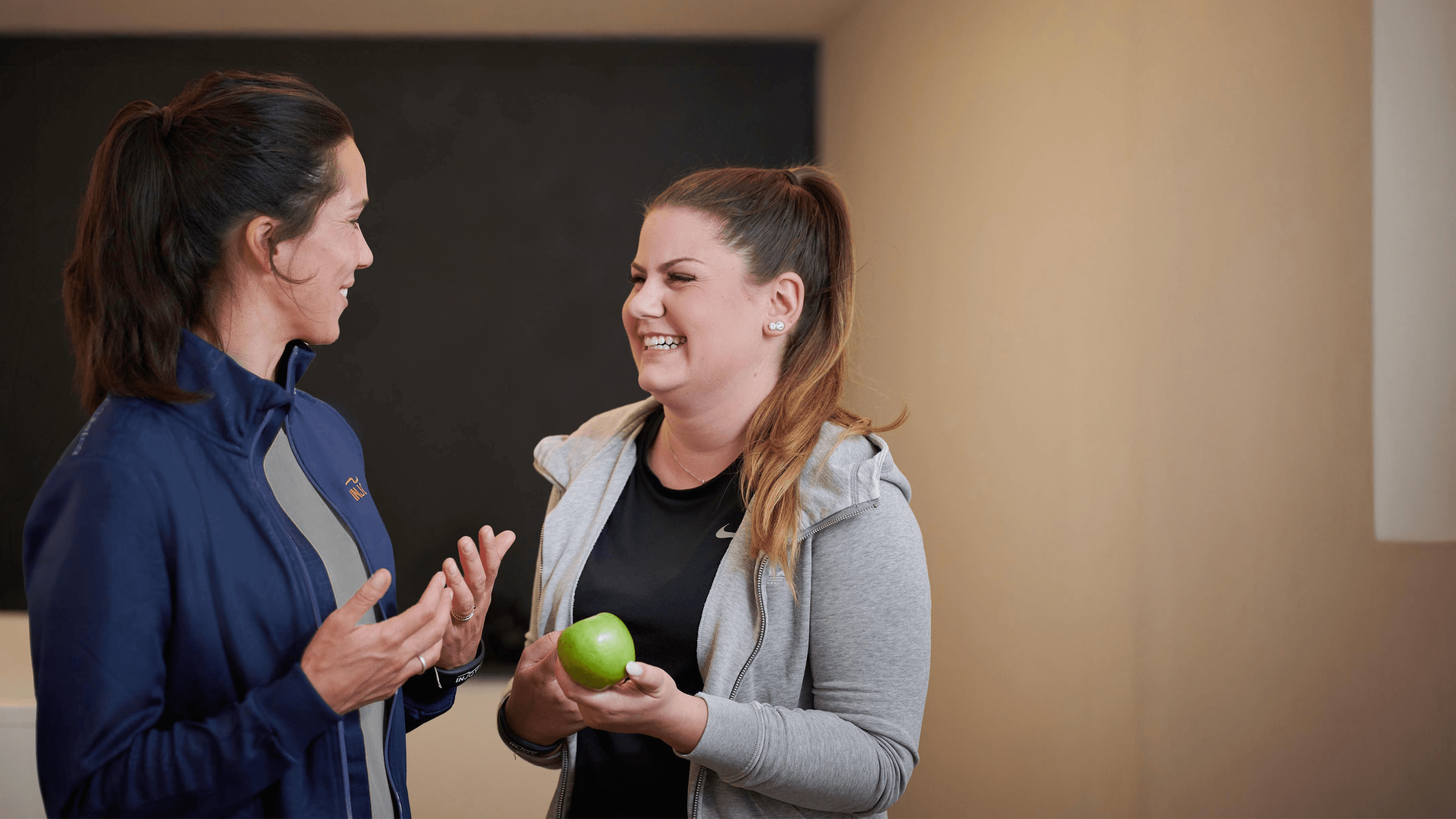 Eine INJOY Trainerin unterhält sich mit einem weiblichen INJOY Fitnessstudio-Mitglied. Die Atmosphäre ist sehr positiv. Das Mitglied grinst die Trainerin an.